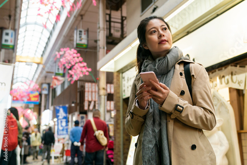 low angle shot of Asian chinese woman visitor looking into space while using gps on phone to look for direction in the arcade food street in Shinsekai area of Osaka Japan