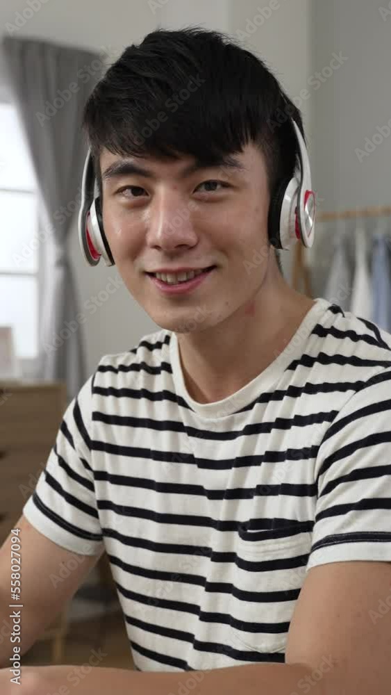 Vertical Screen: portrait of a cheerful asian college boy learning from home with headset is turning head to smile at camera while typing schoolwork on computer in the bedroom. Vertical Screen: portrait of a cheerful asian college boy learning from home with headset is turning head to smile at camera while typing schoolwork on computer in the bedroom.