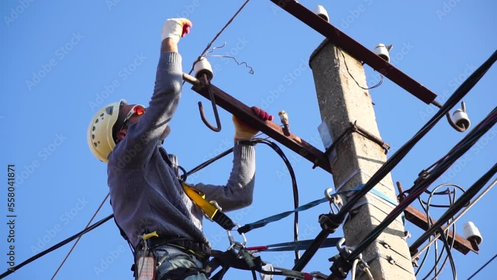 Skilled electrician in helmet fixes wires standing on ladder near high pole against blue sky on summer day backside view. Electrical service and mounting on the pole. Slow motion Skilled electrician in helmet fixes wires standing on ladder near high pole against blue sky on summer day backside view. Electrical service and mounting on the pole. Slow motion