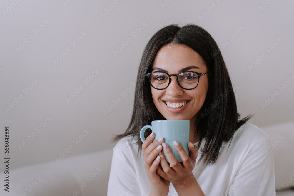 Adorable african american female in glasses and white t-shirt holding cup of coffee, sits at home on sofa looks at camera, broad smiling. Brazilian young woman enjoys holidays. Mock up, awesome girl. Adorable african american female in glasses and white t-shirt holding cup of coffee, sits at home on sofa looks at camera, broad smiling. Brazilian young woman enjoys holidays. Mock up, awesome girl.