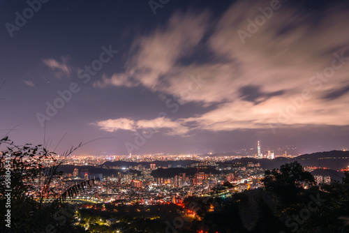 A vibrant cityscape with white clouds moving in the night sky. Night view of the city surrounded by mountains is hazy and dreamy.. Taipei City, Taiwan