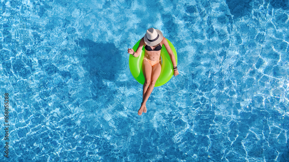 Beautiful woman in hat in swimming pool aerial top view from above, young girl in bikini relaxes and swims on inflatable ring donut and has fun in water on tropical vacation on holiday resort
