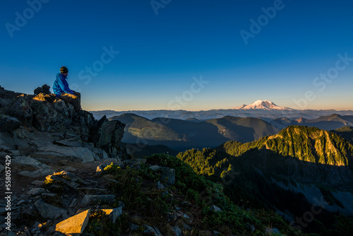 Athletic adventurous male hiker sitting on a ridge on top of a mountain with Mount Rainier in the background during a beautiful sunrise. 