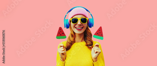 Summer fresh colorful portrait of happy laughing young woman in headphones listening to music with fruit juicy lollipop or ice cream shaped slice of watermelon isolated on pink background