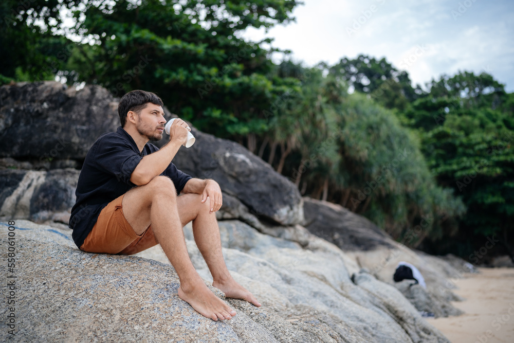 A man drinks a delicious coffee drink on the stones of a tropical beach in the morning in shorts and a black shirt. A man drinks a delicious coffee drink on the stones of a tropical beach in the morning in shorts and a black shirt.