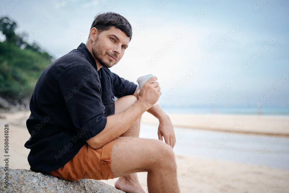 Attractive young man holding a disposable glass with coffee drink on the stones of a tropical beach in the morning and looking at the camera against the sky in summer.
