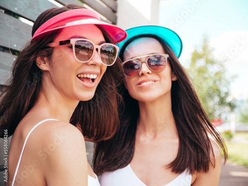 Two young beautiful smiling hipster women posing in the street. Sexy trendy models in summer white tops and colourful skirts. Positive female in sunglasses. Cheerful and happy outdoors. In visor cap © halayalex