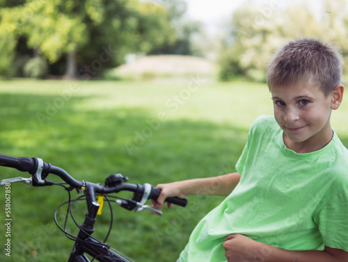  boy of European appearance with short blond hair in a green T-shirt stands with a bicycle in the park against the background of green grass and trees. looks at the camera smiles
