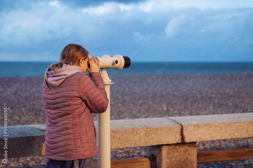 a girl of European appearance with blond hair stands on the embankment looking through binoculars at the ocean. the girl is dressed in a pink jacket and jeans, there is a place for an inscription