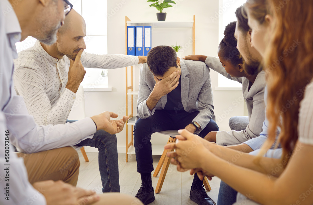 Fototapeta premium Sad upset man at support group therapy session sitting with his head down and the participants touch his shoulders. He is talking his story. Group therapy, psychological help and support concept.