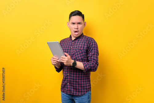 Surprised young Asian man holding digital tablet and looking at camera with open mouth isolated over yellow background