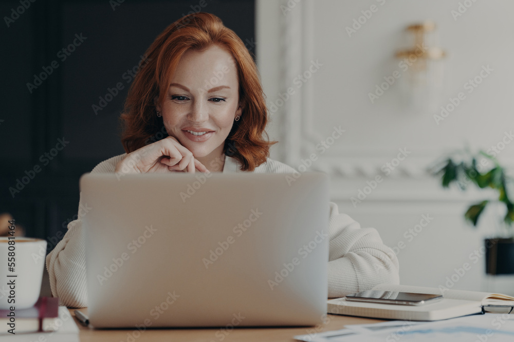 Fototapeta premium Experienced red haired businesswoman reads publication on modern laptop computer, concentrated at screen with pleasant smile, makes research for planning startup, checks mail box, works from home