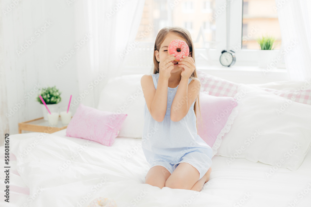 Small pretty female child holds tasty sweet doughnut, poses in bedroom on comfortabled bed, dressed in pyjamas, going to have breakfast, has joy and poses at camera. Chilhood and bedding concept Small pretty female child holds tasty sweet doughnut, poses in bedroom on comfortabled bed, dressed in pyjamas, going to have breakfast, has joy and poses at camera. Chilhood and bedding concept