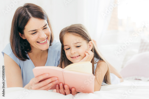 Photography Pretty female mother and her small daughter read interesting book in bedroom, lie on comfortabled bed during morning