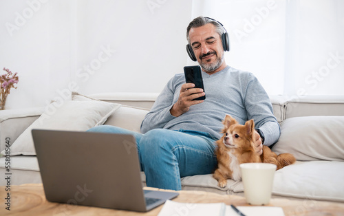 Portrait of senior old caucasian man stay home sit on the sofa  living room listen to music head phone with lovely dog. Happy moment of pure love middle age male playing with little dog in winter © paulaphoto