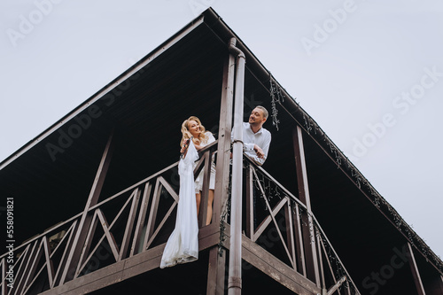 A beautiful curly blonde bride in lingerie, a bathrobe holds a white dress hanging on a hanger, standing on the second floor of a large wooden house, a cottage with a groom. wedding photography. photo