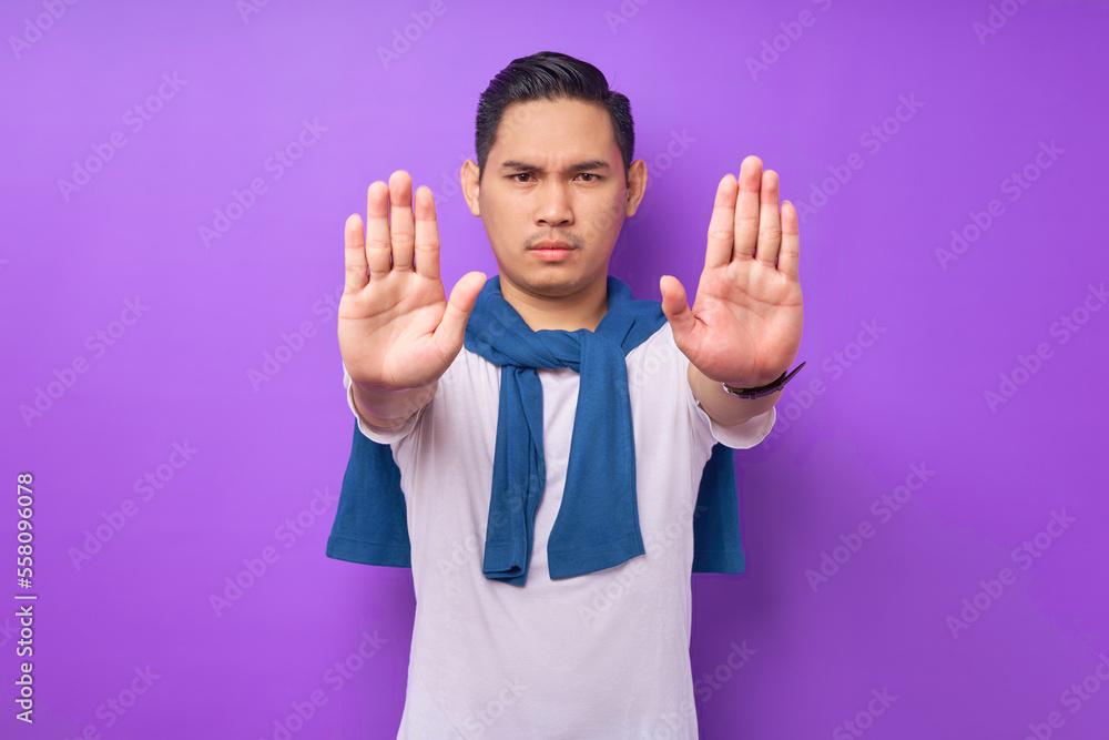 serious strict angry young Asian man wearing white t-shirt showing stop palm gesture refusing isolated over purple background. people lifestyle concept serious strict angry young Asian man wearing white t-shirt showing stop palm gesture refusing isolated over purple background. people lifestyle concept