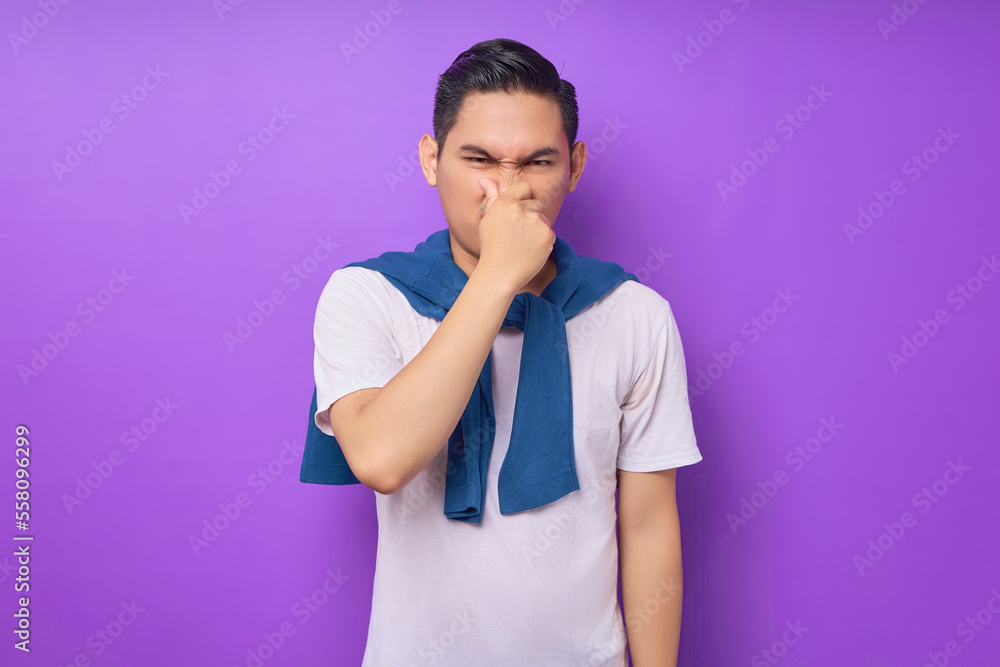 confused young Asian man wearing white t-shirt looking at the camera and covering his nose with a hand isolated over purple background. people lifestyle concept confused young Asian man wearing white t-shirt looking at the camera and covering his nose with a hand isolated over purple background. people lifestyle concept