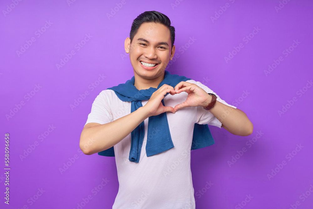 Smiling young Asian man wearing white t-shirt showing shape heart with hands isolated over purple background. people lifestyle concept Smiling young Asian man wearing white t-shirt showing shape heart with hands isolated over purple background. people lifestyle concept