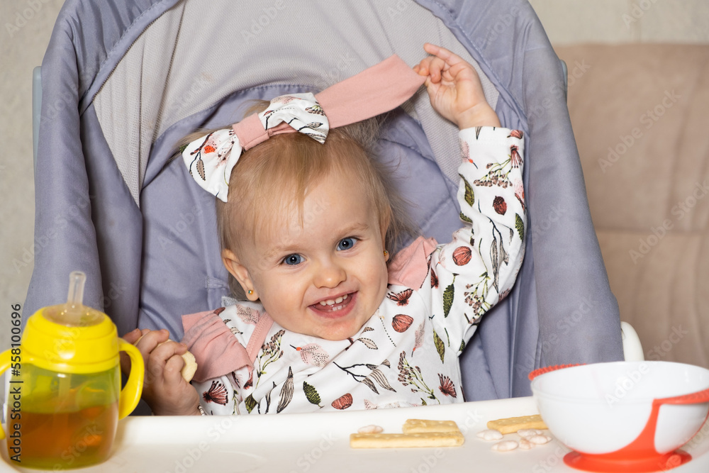 Naklejka premium A happy little girl sits in a high chair with a bottle and a plate and looks into the camera smiling.