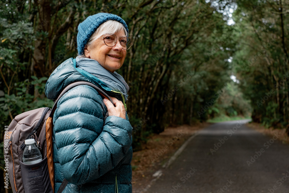 Smiling senior woman enjoying freedom in outdoors walking in a mountain forest. Elderly cheerful woman traveling  in a park holding backpack