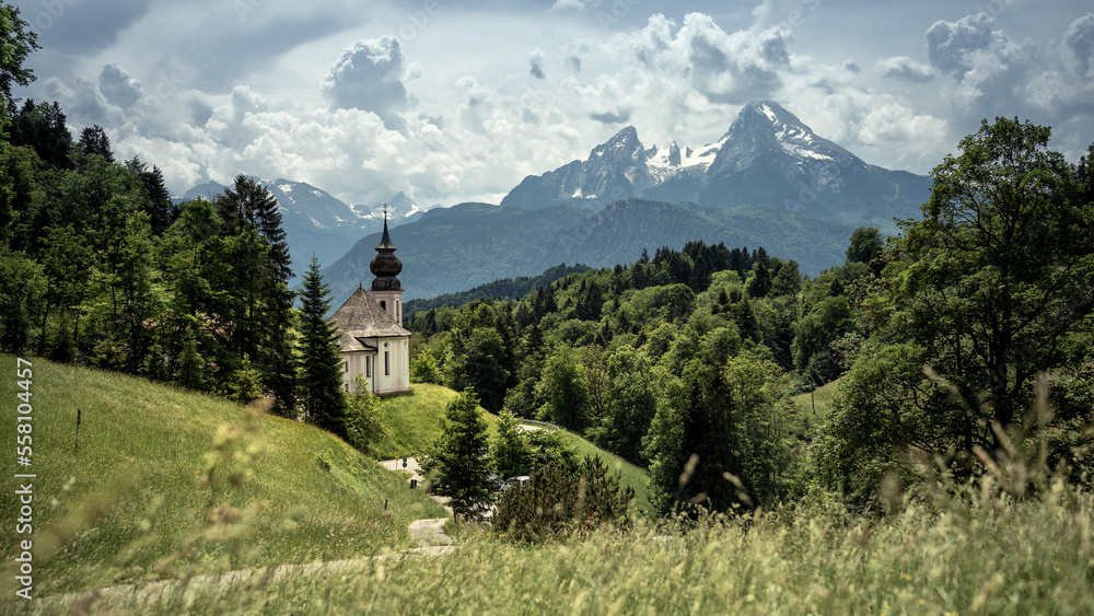 Kirche zwischen grünen Bäumen und Bergpanorama im Hintergrund. In Berchtesgaden mit Watzmann im Sommer.