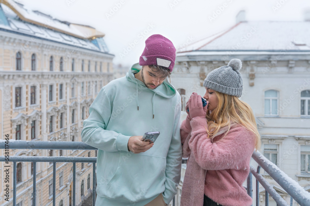 Young couple, she drinks coffee, he looks at cell phone, both on balcony. It snows over the city of Vienna, Austria.