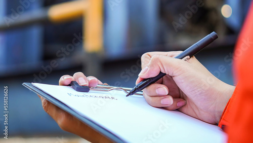 An engineer is writing on paper the to the maintenance detail with a heavy pumping machine as blurred background. Industrial expertise occupation working scene, selective focus at hand's part.