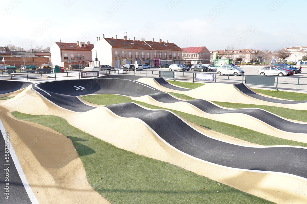 Pump track in black, yellow and with artificial grass; for wheeled vehicles such as bicycles, skates and scooters.