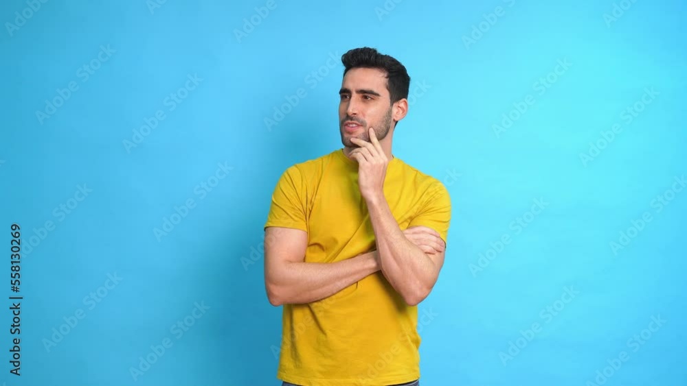 Young bearded male in yellow shirt touching chin and looking at camera while standing against blue background Young bearded male in yellow shirt touching chin and looking at camera while standing against blue background
