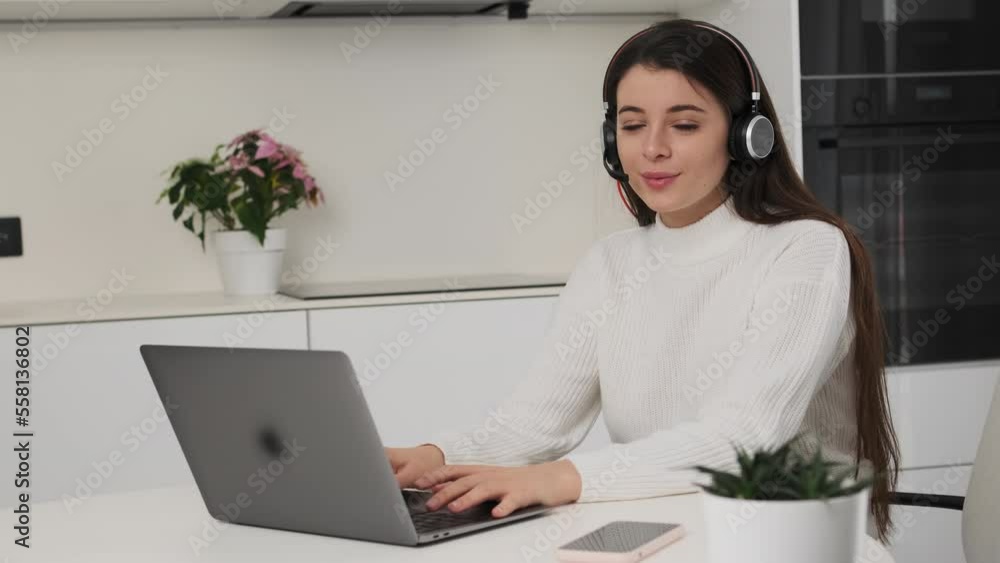 Brunette woman in headset works online on Notebook in kitchen. Young freelancer sits at table turning to look in camera smiling slow motion Brunette woman in headset works online on Notebook in kitchen. Young freelancer sits at table turning to look in camera smiling slow motion