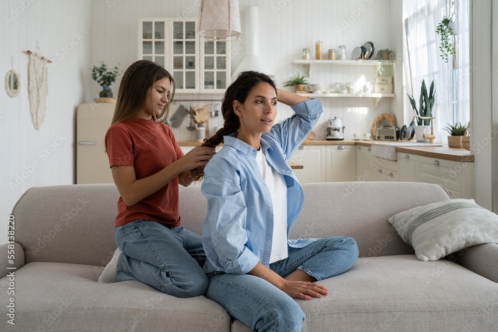 Teenage girl daughter braiding hair and making hairstyle for her attractive charming mother while enjoying time together at home, sitting on sofa in living room. Spending weekend with mom Teenage girl daughter braiding hair and making hairstyle for her attractive charming mother while enjoying time together at home, sitting on sofa in living room. Spending weekend with mom