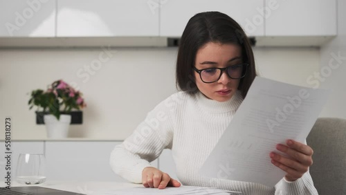 Young employee in black-rimmed glasses checks project papers sitting at table near laptop. Focused brunette woman enjoys working at home photo
