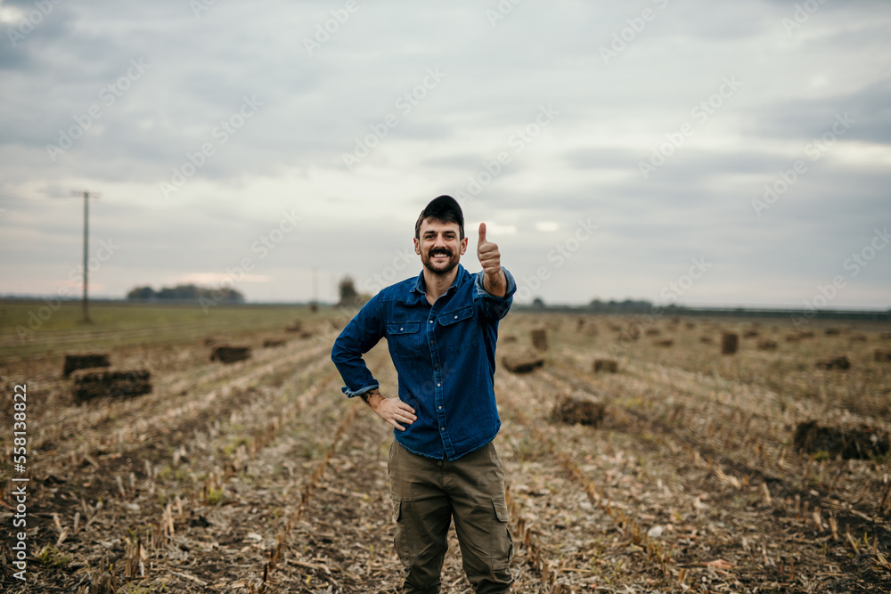 Proud worker man in a work outfit standing in his field after a good and productive agricultural season. Hay bales behind him.
