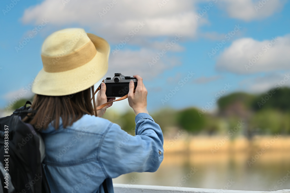 Obraz premium Rear view of woman traveller with backpack taking photo with digital camera while standing on a bridge over a canal