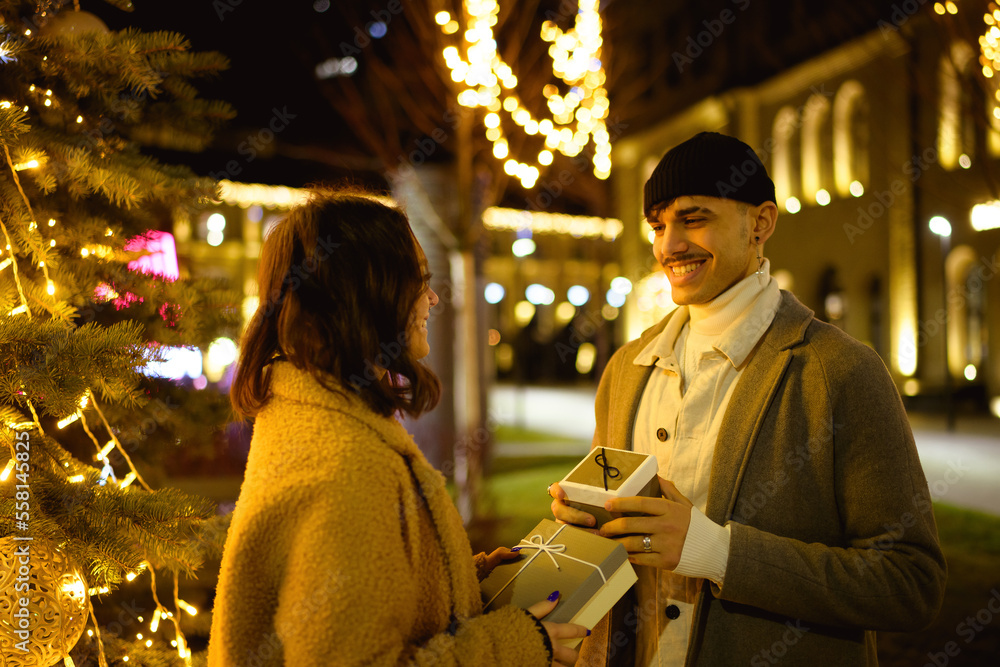 Young romantic couple holding gifts near blurred lights on urban street in evening during New Year