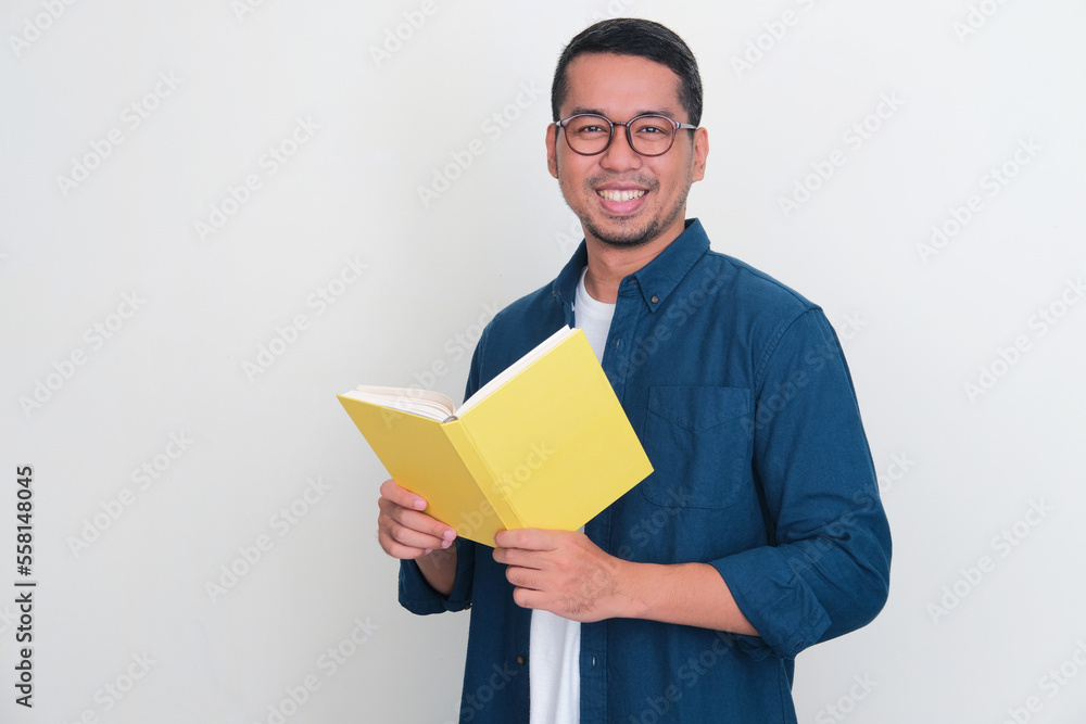 Adult Asian man smiling friendly at the camera while holding a book Adult Asian man smiling friendly at the camera while holding a book