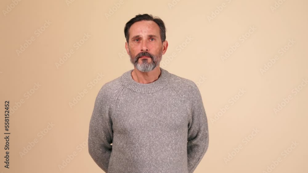Concentrated adult bearded man in grey sweater showing silence gesture at the camera in the beige studio Concentrated adult bearded man in grey sweater showing silence gesture at the camera in the beige studio