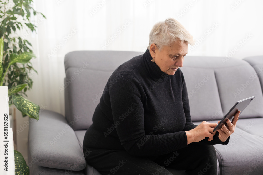 Smiling middle-aged Caucasian woman sit on couch in living room browsing wireless Internet on tablet, happy modern senior female relax on sofa at home using pad device, elderly technology concept Smiling middle-aged Caucasian woman sit on couch in living room browsing wireless Internet on tablet, happy modern senior female relax on sofa at home using pad device, elderly technology concept
