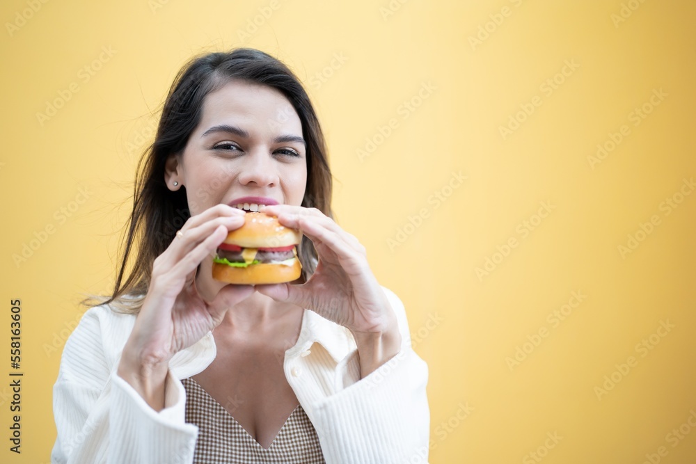 Cheat Meal Pretty Lady Eating Hamburger Junk Food. Cheerful Woman Smelling Burger Standing Over Yellow Background. Studio Shot Cheat Meal Pretty Lady Eating Hamburger Junk Food. Cheerful Woman Smelling Burger Standing Over Yellow Background. Studio Shot