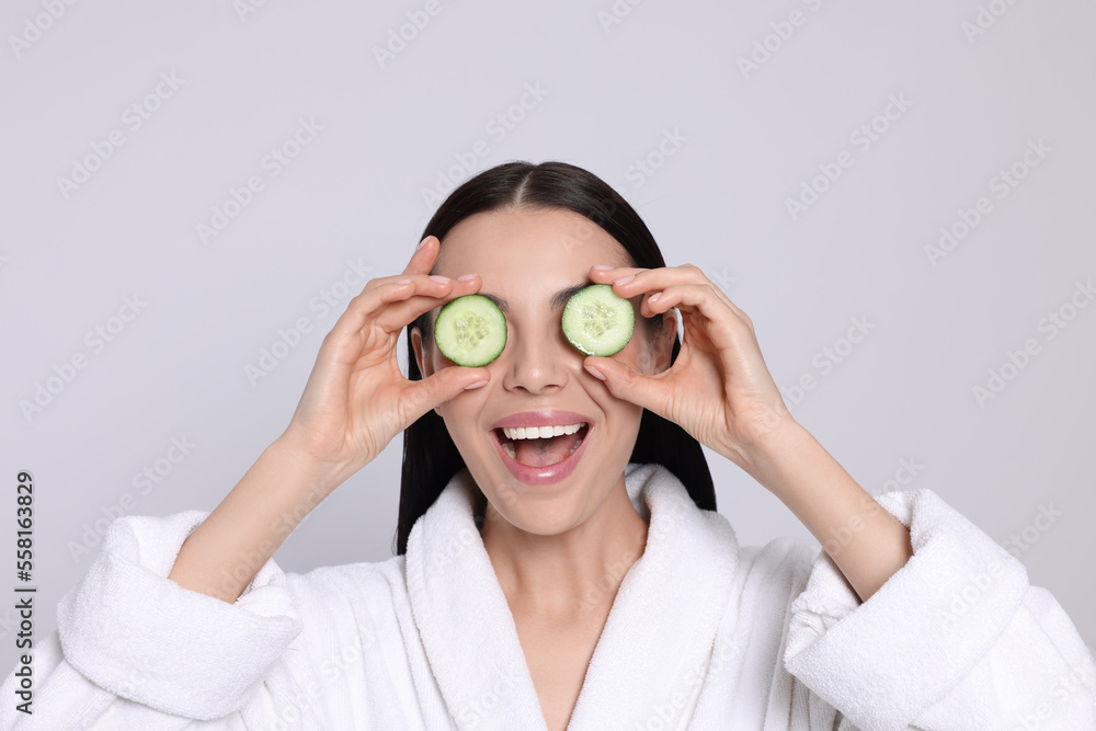 Woman in bathrobe holding pieces of cucumber on light grey background. Spa treatment Woman in bathrobe holding pieces of cucumber on light grey background. Spa treatment