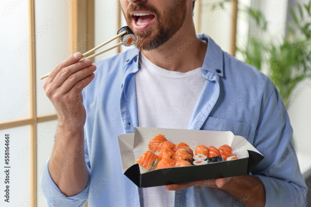 Handsome man eating sushi rolls with chopsticks indoors, closeup