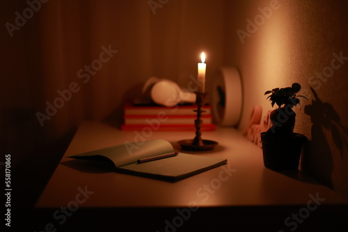 In the dark room, burning candle in candlestick is lighting on the work desk where the paper notebook with pen. Studying and working at home at blackout. Problem with electricity