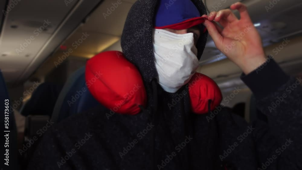 Male tourist in medical mask and eye bandage is trying to relax in airplane salon with neck pillow around neck. Tired man prepares for long flight with comfortable devices. Flying on jet on quarantine