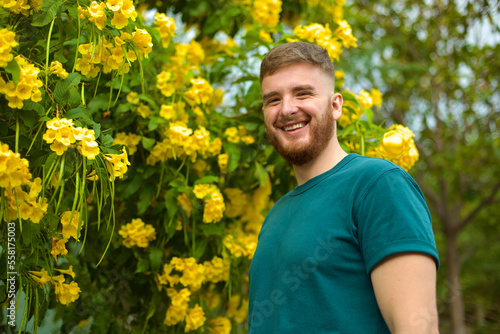 Portrait of happy handsome bearded guy, young positive man with beard is smelling beautiful yellow flowers in the garden, smiling, enjoying spring or summer day, breathing deep deeply fresh air © Евгений Шемякин