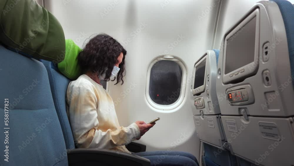 Profile view of caucasian female passenger sitting in plane in medical mask and using smartphone. Woman is travelling by jet during pandemic and surfing the internet on mobile waiting for the flight Profile view of caucasian female passenger sitting in plane in medical mask and using smartphone. Woman is travelling by jet during pandemic and surfing the internet on mobile waiting for the flight