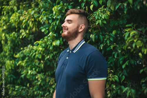 Portrait of a beautiful young bearded man with beard standing on an Ivy wall or in greenery, smiling against from plant, guy relaxing outdoors at sunny summer day. Love nature, eco life