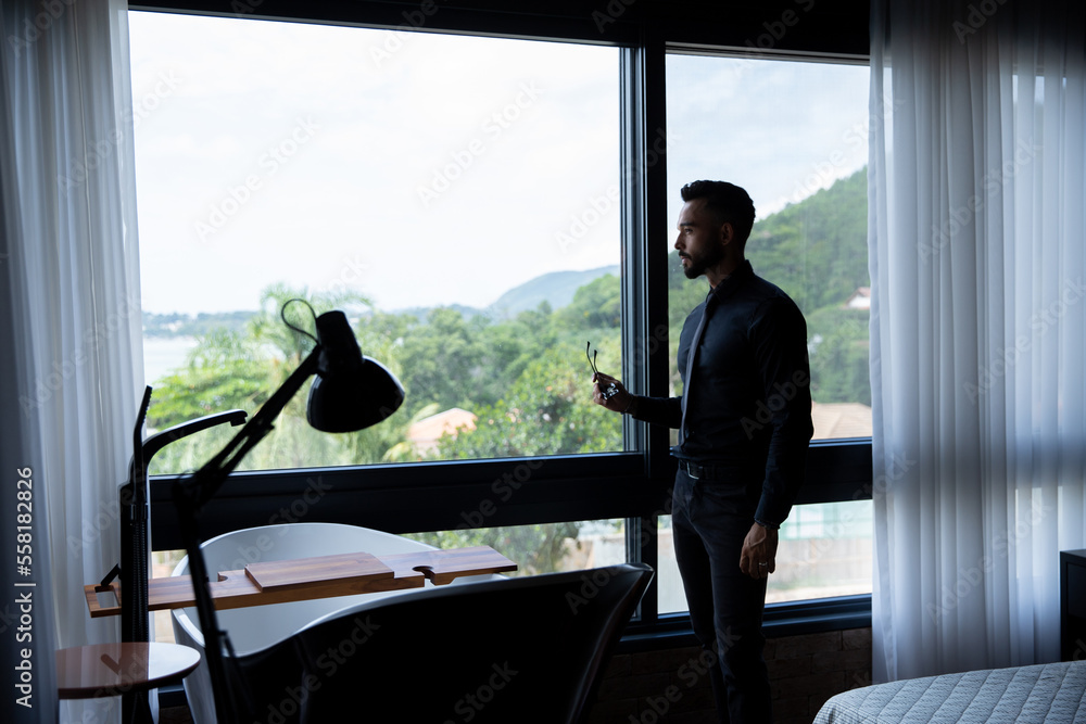 man wearing business suit standing in front of a window, young businessman in his luxury room looking out the window wearing blouse and black tie