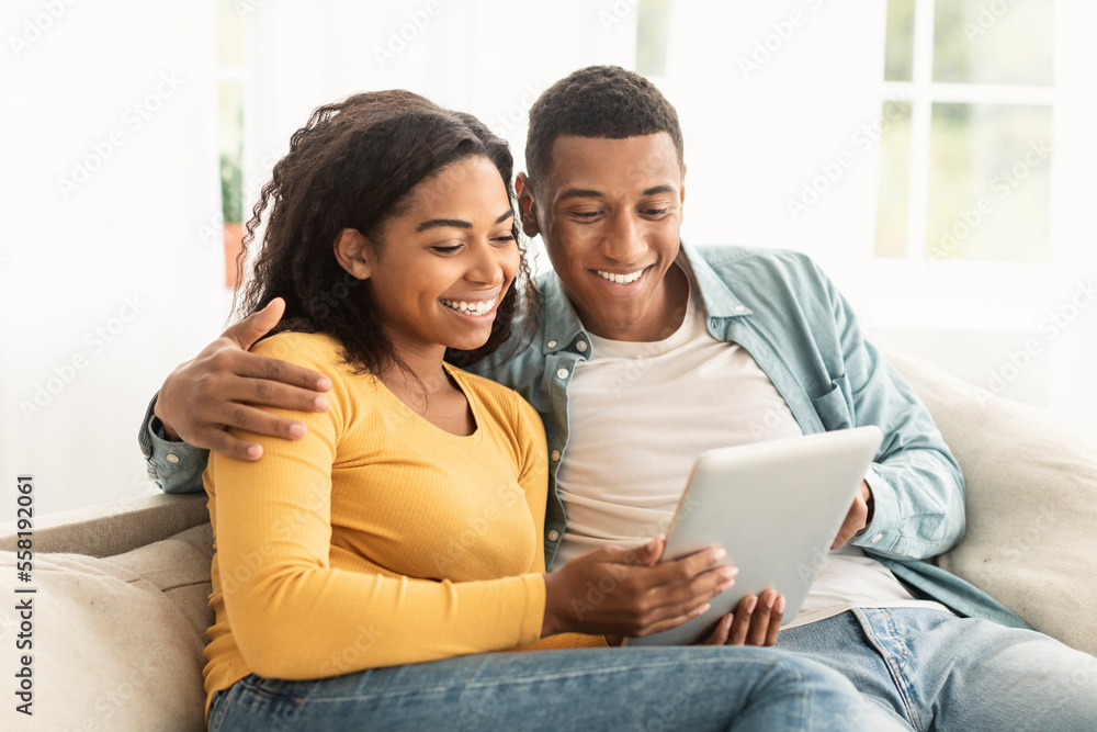 Smiling millennial african american husband and wife surfing in internet, have meeting, watch video on tablet