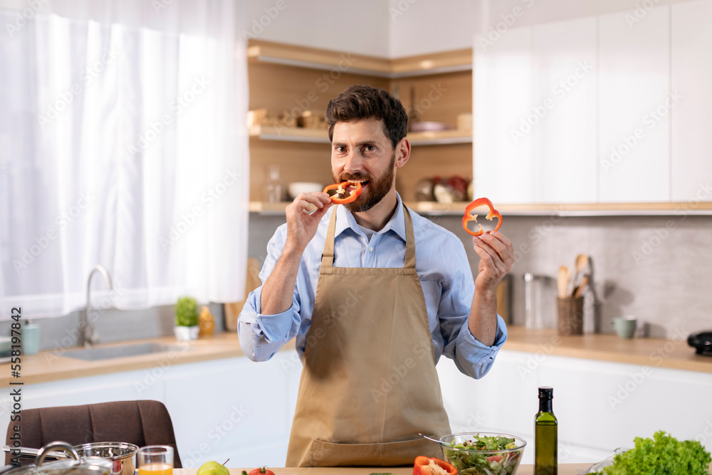 Happy millennial caucasian male in apron prepares food, bites piece of pepper, enjoys cooking at table